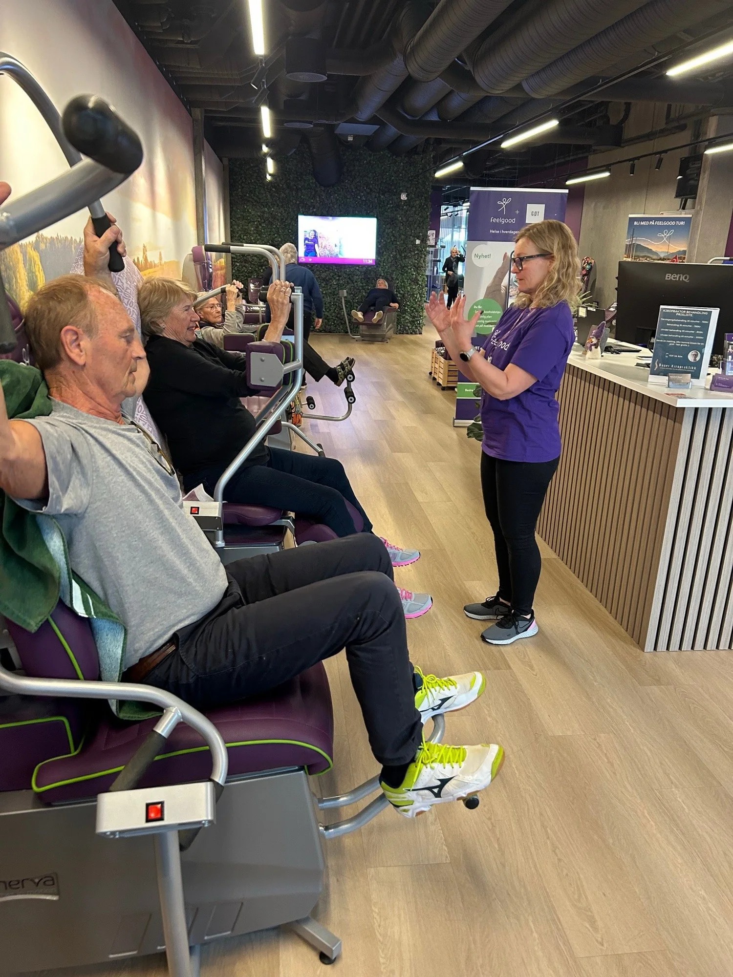 Medlemmer i sirkeltrening hos Feelgood Heggedal, med Bauer Kiropraktikk sin prisliste og «Bli med på Feelgood tur»-banner i bakgrunnen
